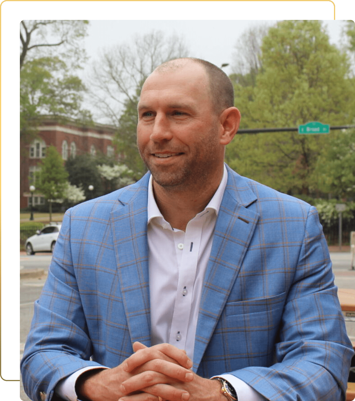 Well-dressed man in blue plaid jacket smiles confidently outside brick building on tree-lined street